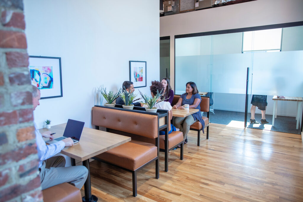 A modern office space with wooden floors and frosted glass partitions. Four people sit at booth tables with laptops and coffee, surrounded by plants, creating a collaborative and relaxed atmosphere.