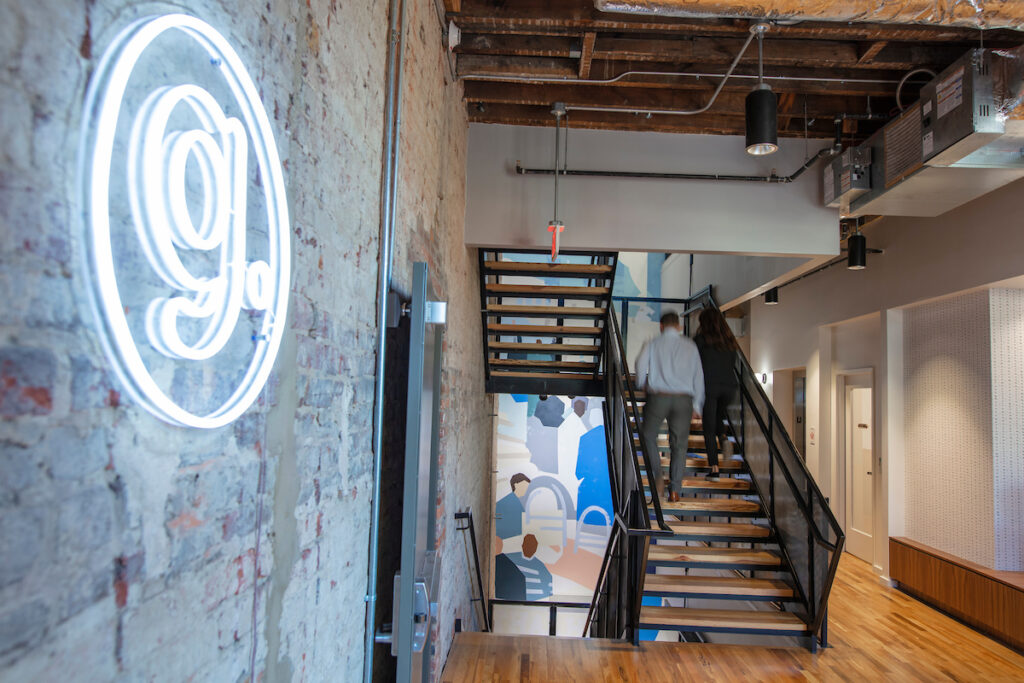 Modern office interior with exposed brick walls, a glowing "g." sign, a staircase with metal railings, wooden flooring, and a colorful wall mural.