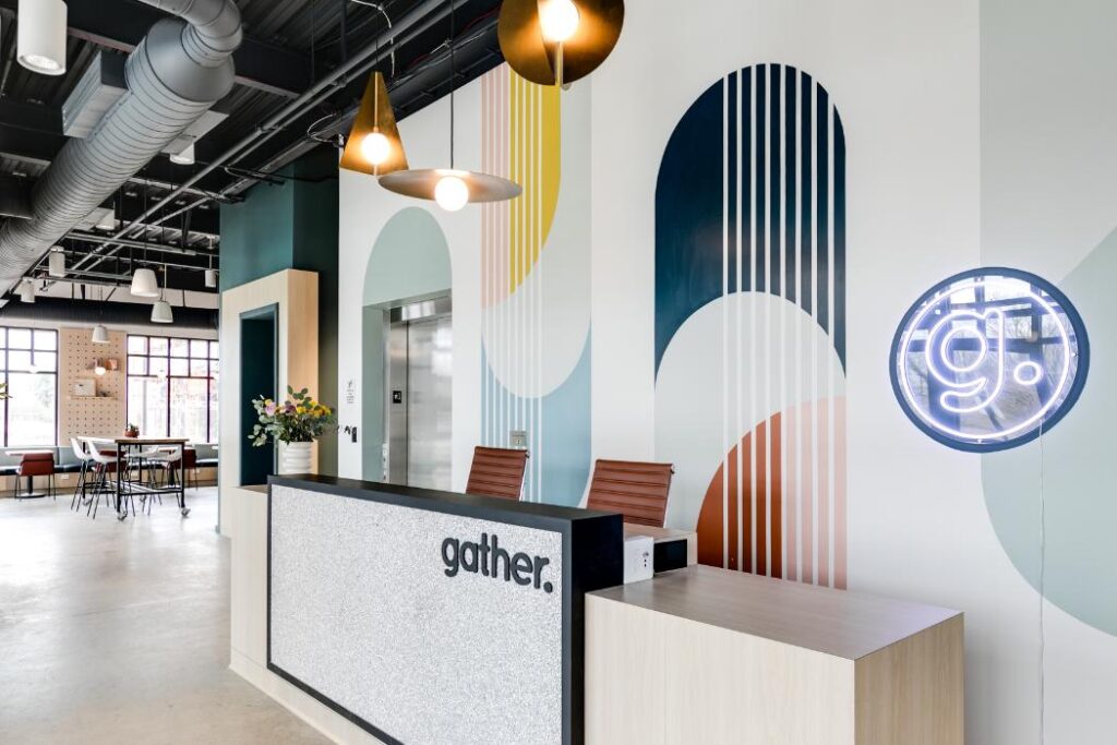 Modern office space with a reception desk labeled "gather." Colorful wall art with abstract shapes, overhead lights, and a glowing round sign. Calm and inviting ambiance.