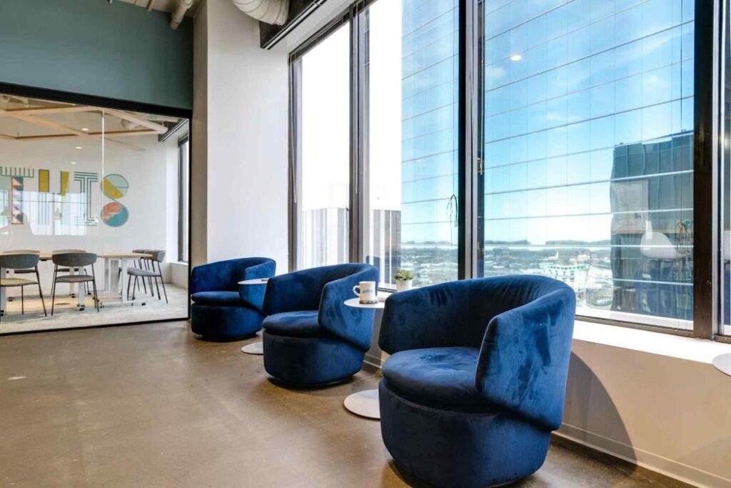 Modern office lounge with three blue velvet chairs and small round tables by large windows. A glass-walled meeting room is visible on the left.