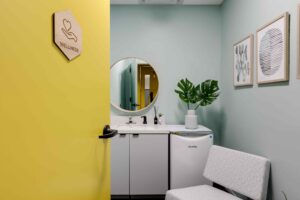 Wellness room Gather Norfolk with a yellow door partially open, revealing a calm space. Inside are a sink, round mirror, potted plant, small fridge, and abstract wall art.