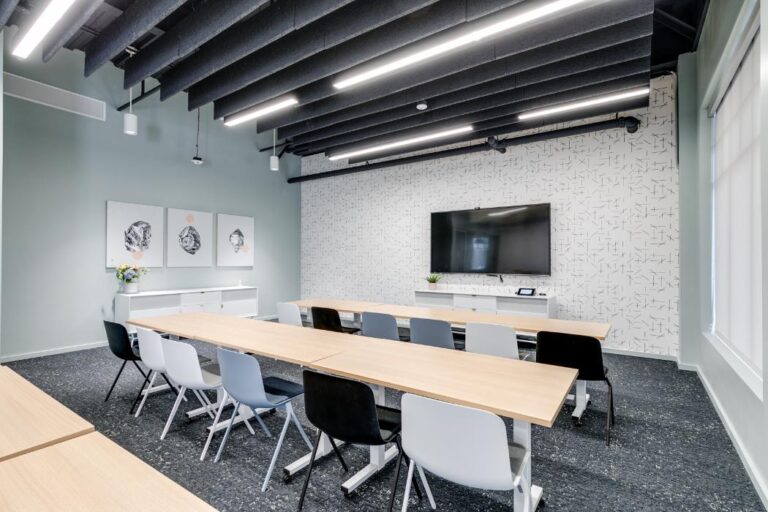 Modern conference room with long wooden tables, black and white chairs, wall art, large flat-screen TV, and patterned wallpaper. Bright, minimalistic design.