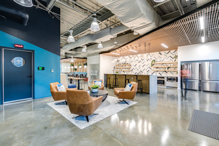 Modern office lounge Newport News, VA, with tan leather chairs around a small table on a rug. Open kitchen area in the background with bar stools and patterned backsplash.