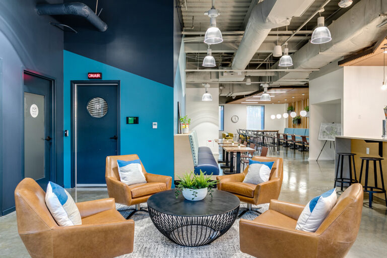 Newport News, VA, Modern office lounge with four tan leather chairs around a black round table, plant centerpiece. Blue and white walls, industrial ceiling, inviting ambiance.