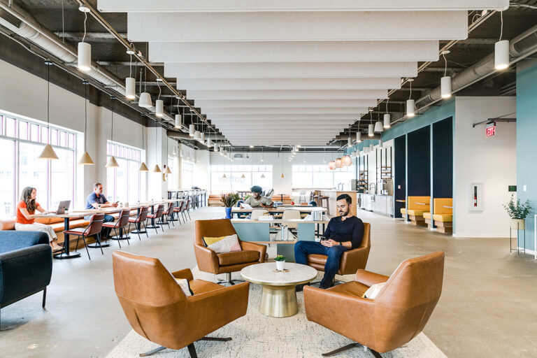 Spacious, modern office lounge Virginia Beach, VA, with brown leather chairs and a round table in the foreground. People work and relax in bright, airy surroundings.
