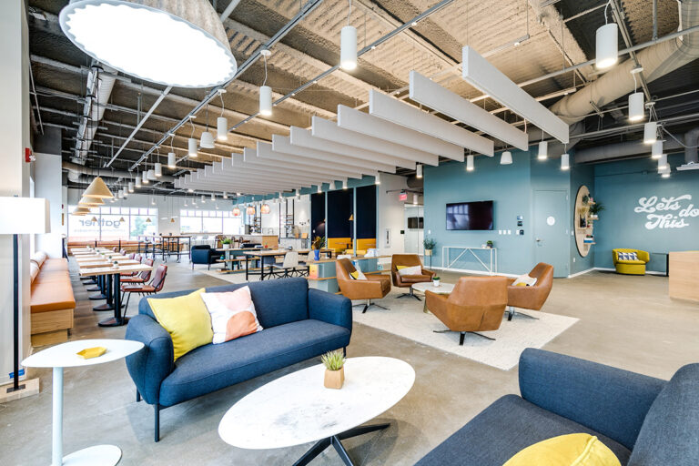 Modern coworking space in Virginia Beach, VA with blue sofas, brown armchairs, and accent pillows under a ceiling with white panels. Bright, inviting, and collaborative atmosphere.