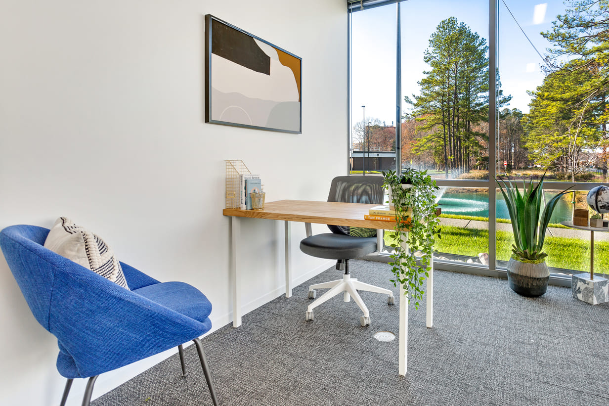 Private office at Gather with wood desk, ergonomic chair, blue accent chair, and floor-to-ceiling window overlooking trees and water.