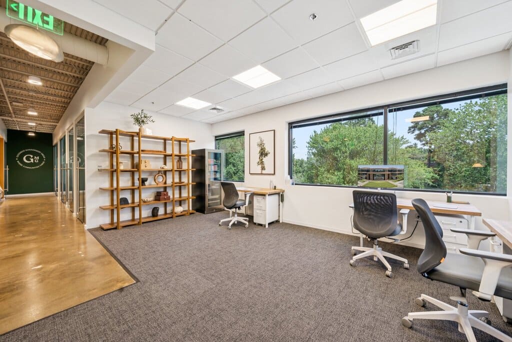 Private office suite at Gather coworking with two desks and ergonomic chairs, large windows with tree views, and open shelving near a glass hallway.