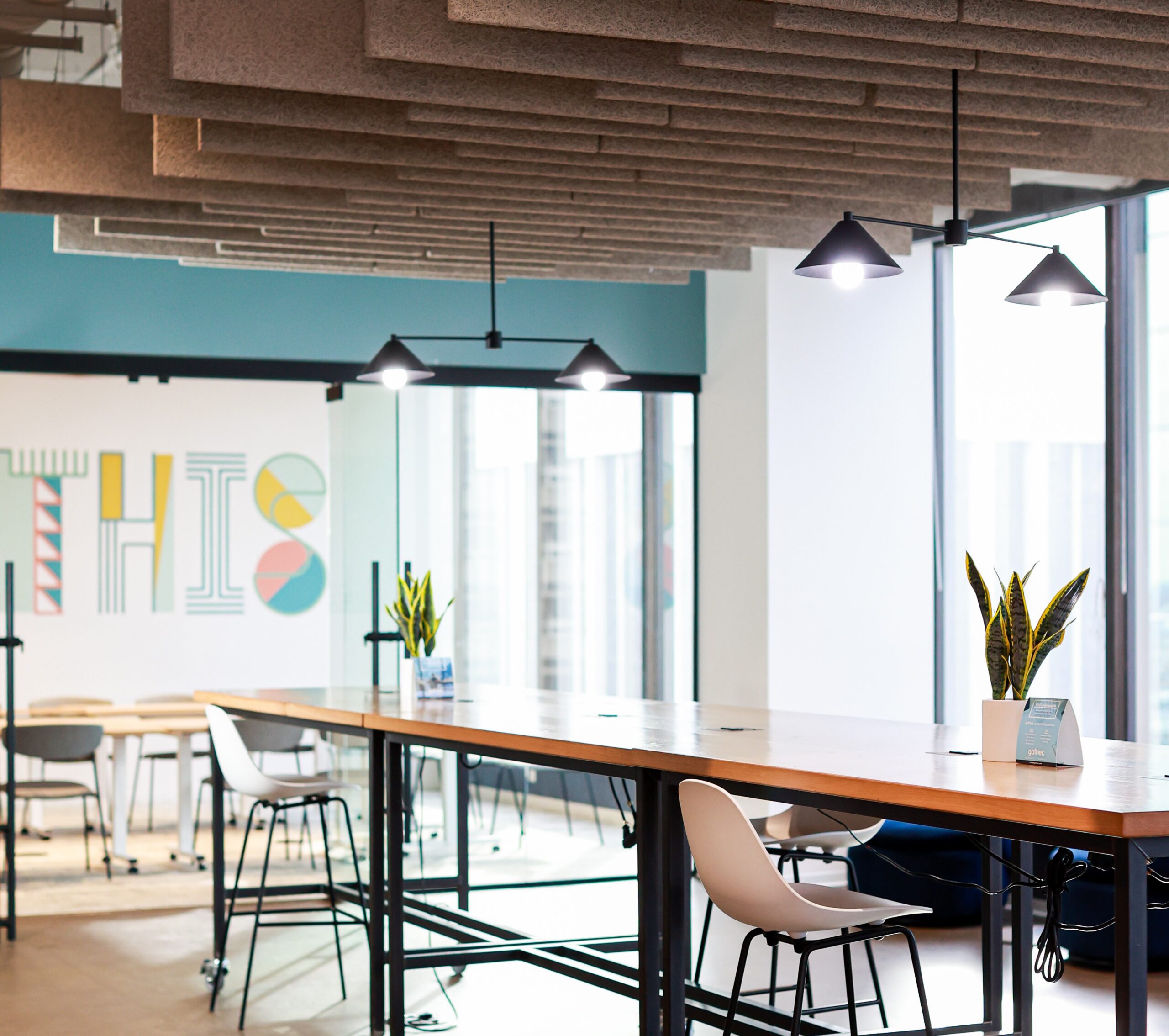 Modern coworking space interior at Gather Norfolk featuring standing height tables, acoustic ceiling panels, glass-walled conference room, and colorful branded wall art.