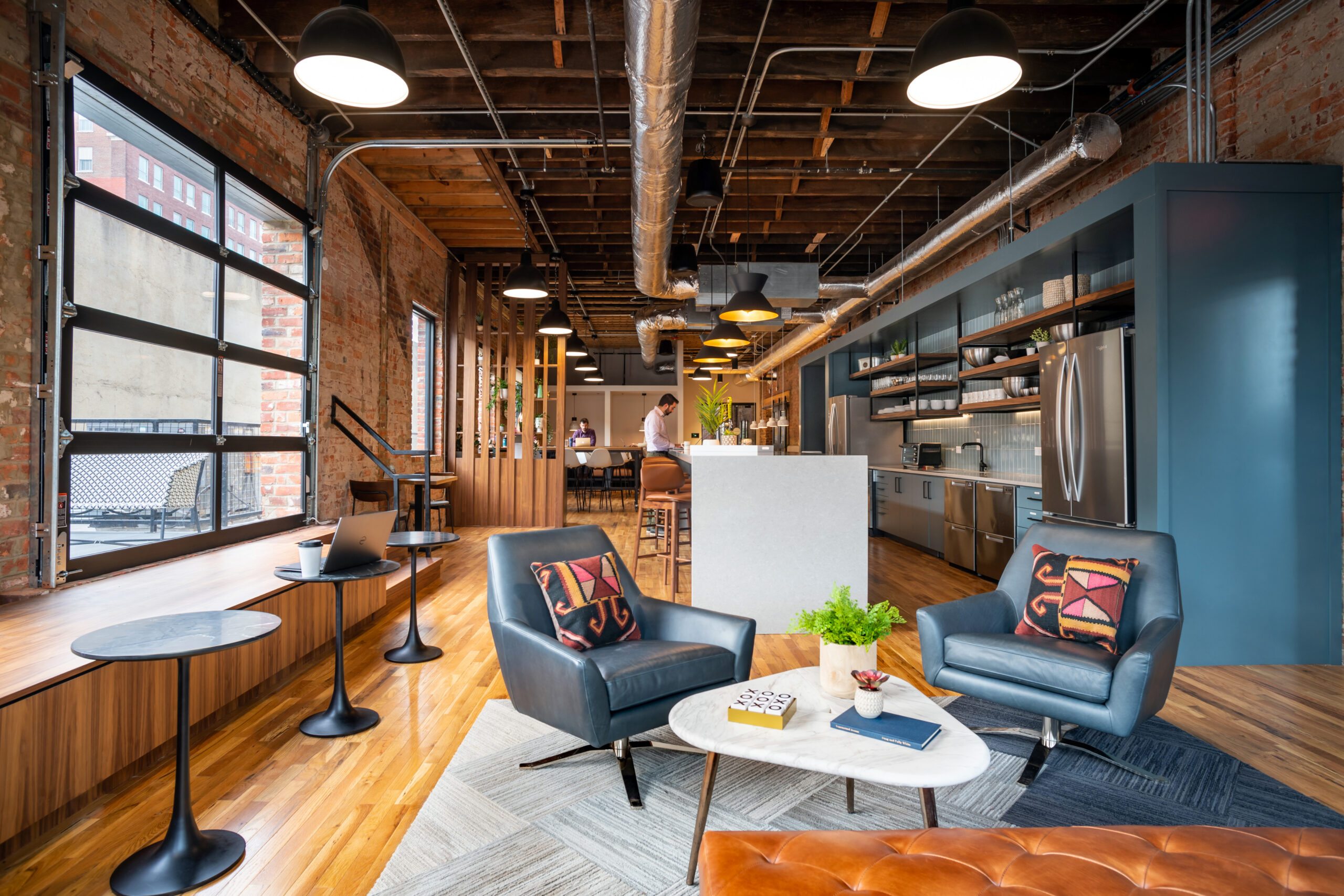 Professional coworking space interior at Gather' Hellos Richmond Arts District location featuring exposed brick walls, natural light from large windows, modern lounge seating, standing desk workspace, and fully equipped kitchen