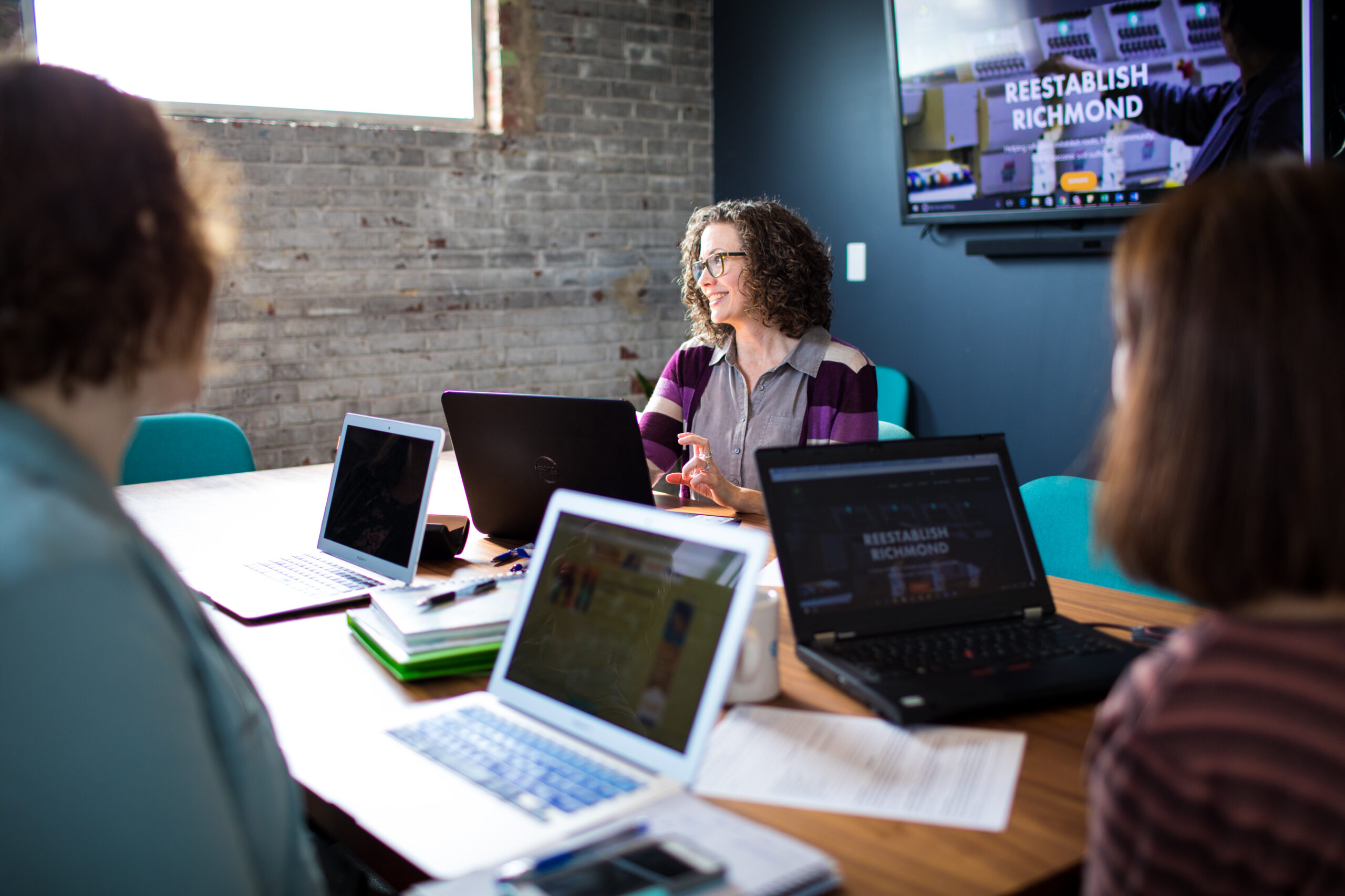 Team meeting in progress at Gather Scott' Hellos Addition conference room with exposed brick walls, mounted monitor, and collaborative workspace setup