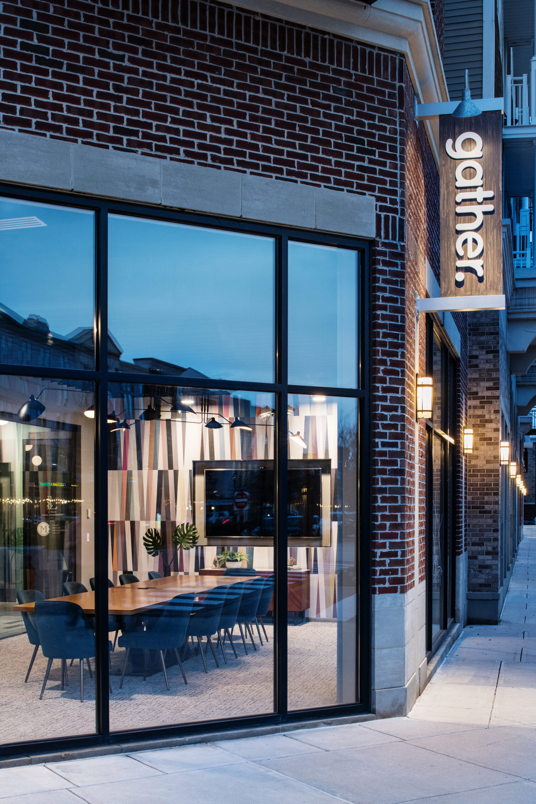 Evening exterior view of Gather Short Pump coworking space featuring modern brick architecture, large windows, and illuminated conference room