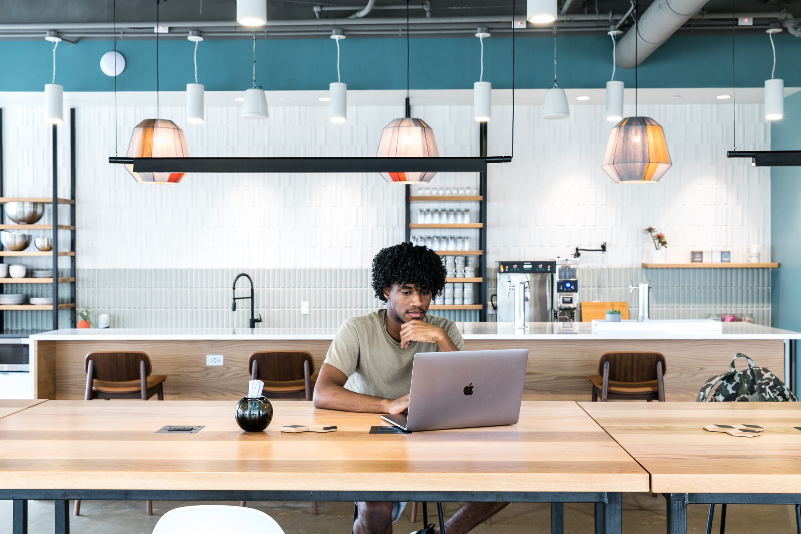 Professional working at laptop in Gather Virginia Beach coworking space with modern kitchen, designer pendant lighting, and collaborative workspace.